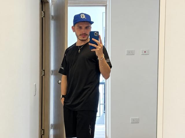 LianFerrer stands in a doorway, wearing a blue Yankees cap, black shirt, and black pants, taking a selfie with a blue phone, showing a slight mustache and bracelet.