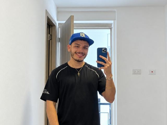 LianFerrer with a blue cap, black t-shirt, and cross necklace, holding a phone, smiling, standing in a doorway, white walls, and wooden door in the background.