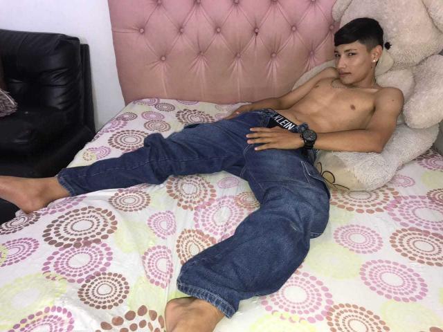 SantiDiazy, sem camisa, com um corpo tonificado, esguio e atraente, vestindo jeans azuis, deitado em uma cama estampada, com uma cabeceira rosa com detalhes em relevo e um grande brinquedo de pelúcia..
