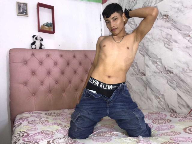 SantiDiazy, sem camisa, com um corpo esguio e tonificado, usando roupa íntima preta e calças jeans abertas, ajoelhado em uma cama, braço direito levantado, braço esquerdo atrás da cabeça, olhando para a câmera..