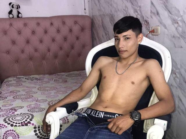 SantiDiazy, sin camisa, con un cuerpo delgado y tonificado, vistiendo jeans y una cadena, sentado en una silla, mirando a la cámara, con un reloj negro y un collar..