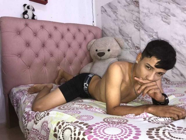 SantiDiazy é um jovem latino de físico esguio, com cabelo preto curto, deitado sem camisa em uma cama rosa, vestindo shorts de boxe pretos, com um dedo na boca e um ursinho de pelúcia..