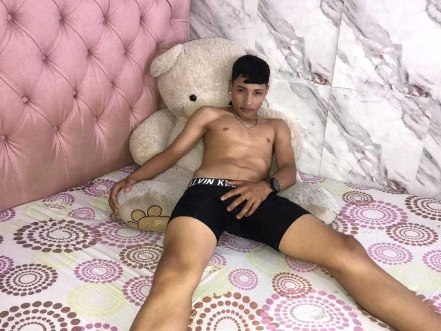 SantiDiazy está sin camisa, vistiendo ropa interior negra, y está acostado en una cama con un cabecero rosa acolchado y un gran oso de peluche blanco. SantiDiazy tiene una complexión delgada y un pene pequeño..