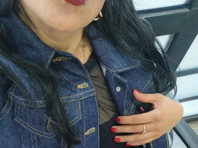 ZarayFeet met een donkere denimjas, rode nagellak en een ring, zwart haar en rode lippenstift, hand op de borst, met een beetje zwart top te zien, zittend op een zwarte stoel..