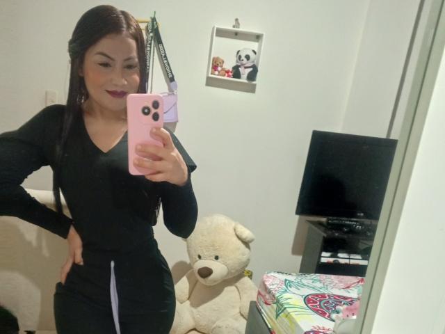 NinnaBallack with dark red lipstick, wearing a black long-sleeve top and black pants, taking a mirror selfie with a pink phone, standing next to a white stuffed dog.