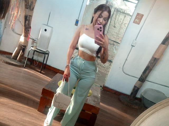 NinnaBallack with long brown hair, white crop top, showing her tits, and light green pants, taking a mirror selfie with a pink phone, holding a white purse.