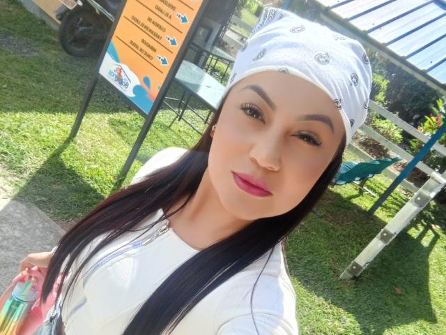 NinnaBallack with white bandana, pink lipstick, black eyebrows, long black hair, white shirt, holding pink and green bag, looking at camera, sunny grassy background.