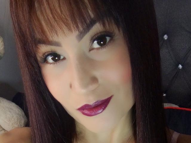 NinnaBallack's face with dark brown straight hair, bangs, brown eyes, black eyeliner, purple lipstick, and a subtle smile. She has a light skin tone and a cute, sexy look.