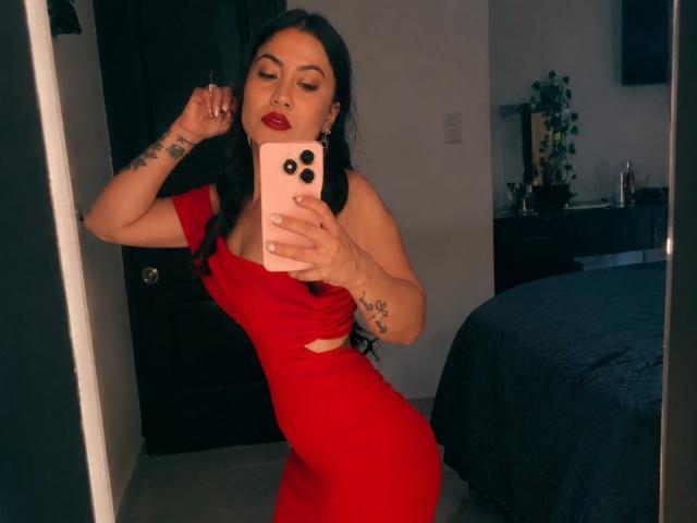 NinnaBallack in a red dress, showing her curvy ass and small tits, taking a mirror selfie with a pink phone, black hair, red lipstick, tattoos on her arm.