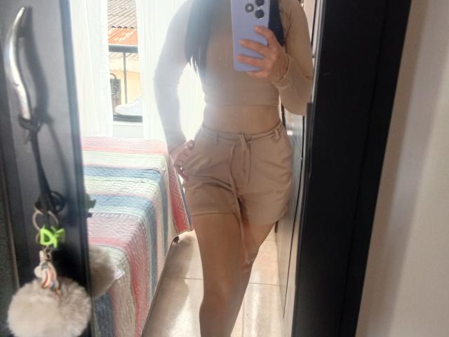 NinnaBallack in beige crop top and matching shorts, holding a phone in a mirror selfie, showing her ass and a hand on her hip, standing in a sunlit room.
