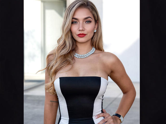 Sofie White, con i capelli biondi, il rossetto rosso, l'abito nero e bianco senza spalline, la collana di diamanti e il braccialetto, in piedi con le mani sui fianchi, che mostra il seno e un tatuaggio sul braccio..