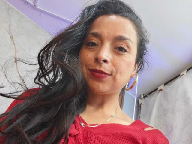 StangelarRivera with wavy black hair, red lipstick, and red top, smiling slightly, wearing gold hoop earrings, and a silver necklace, looking at the camera.