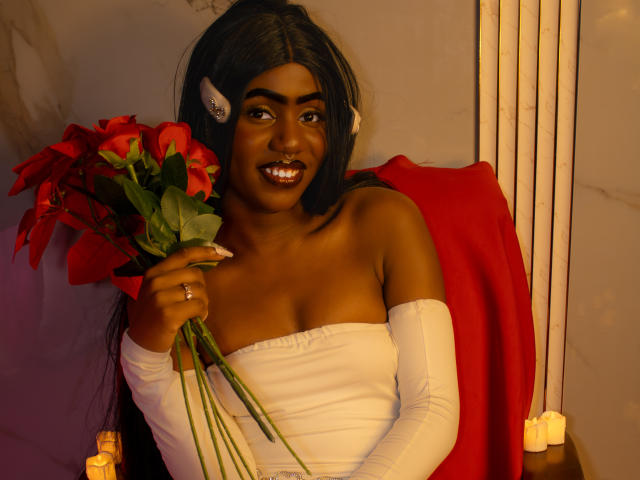 HaryAndKimmy, dark-skinned woman with long black hair, holding red poinsettias, wearing white strapless top and arm sleeves, red cloth behind, smiling with white teeth, ear clip.