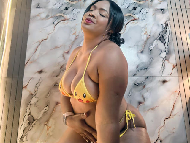 AliceBooty in a yellow bikini, showing off her big tits and curvy ass, with a hand on her pussy, smiling with eyes closed, wearing a pink smartwatch.