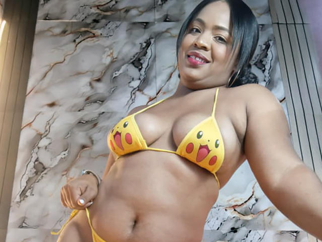 AliceBooty with dark skin, wearing a yellow Pikachu bikini, showing her big tits and small yellow panties, smiling with pink lipstick, black hair, and a curvy, sexy body.