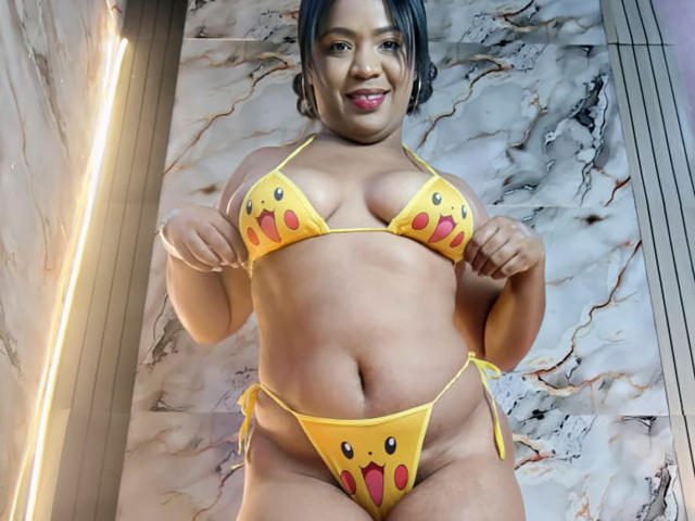 AliceBooty with cute yellow bikini featuring red mouth emojis, smiling, showing her big tits and fat ass, standing in front of marble wall, looking down at the camera.