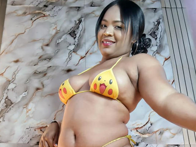 AliceBooty wearing a yellow Pikachu bikini, showing her tits and ass, smiling with dark hair in a ponytail, standing in front of a marble wall.