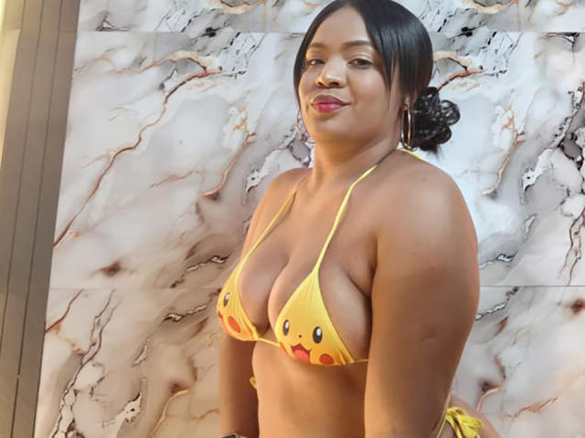 AliceBooty in a yellow bikini with colorful dots, showing her big boobs, with a confident smile, and her black hair in a bun. She has a cute, sexy look.