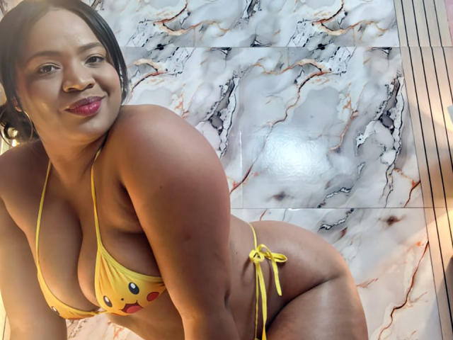AliceBooty, curvy with big boobs, wearing a yellow string bikini with a cute face design, leans forward smiling, showing her yellow tied side-bikini and dark hair.