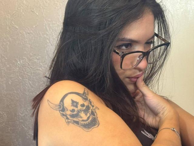 KatarinaNovak with glasses, dark hair, and a skull tattoo with horns on her shoulder, biting her finger, wearing a brown top, in a photo.