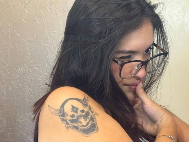 KatarinaNovak with dark, messy hair, glasses, and a smirk, showing a skull tattoo on her right shoulder, touching her chin. Her hand's finger rests on her lip.