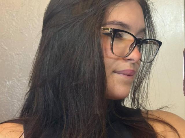 KatarinaNovak with long dark brown hair, black tortoiseshell glasses, and a subtle smile, wearing a black sleeveless top, showing a slight hint of her tits.