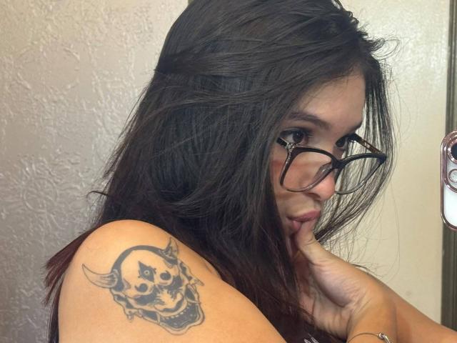 KatarinaNovak with dark hair, glasses, and a skull tattoo on her shoulder, biting her finger, showing off her tits.