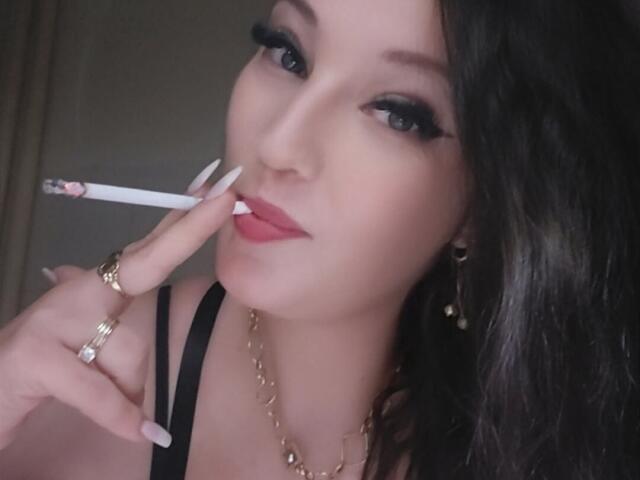 VanessaSquirtt with black hair, wearing a black top, gold chain, and rings, holding a cigarette to her red lips, looking at the camera with black eyeliner.