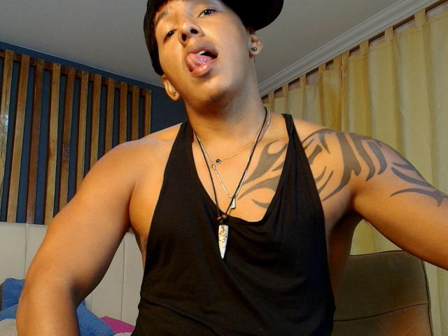 Chico Yaoi con piel oscura, camiseta negra sin mangas y tatuajes, sacando la lengua, usando una gorra negra y un collar, brazos musculosos, cortinas amarillas en el fondo..