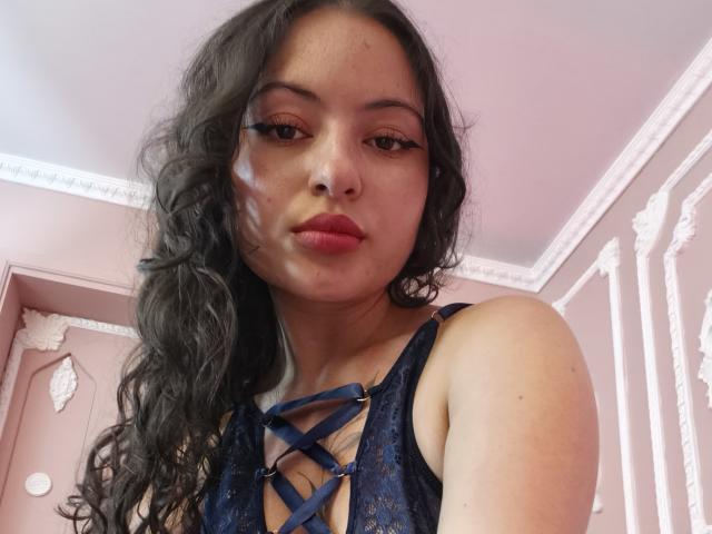 MiiaBennet with wavy dark hair, wearing a blue lace-up top, has a white liquid on her face, looking at the camera with pink lipstick, in a pink room.