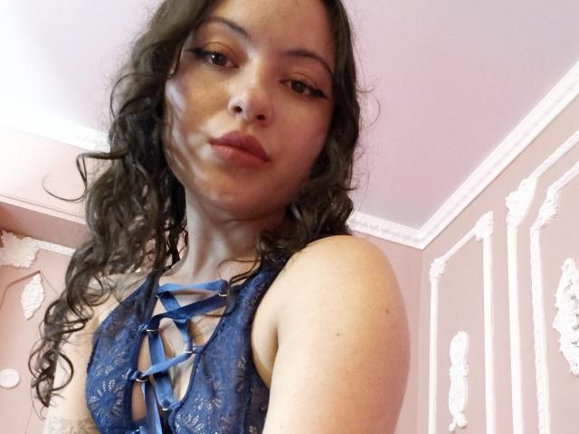 MiiaBennet with curly brown hair, wearing a blue lace lingerie top with blue ribbon details, pouting slightly, looking at the camera. Her tits are partially visible through the lace.