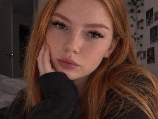 ClarineSoulia with red hair, freckles, and long eyelashes, resting her hand on her cheek, has a pierced lower lip and is wearing a black long-sleeve shirt.