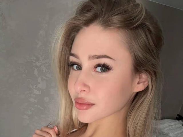 Het gezicht van Milagros Edmisten, met blond haar, blauwe ogen, volle, roze lippen, subtiele make-up en lange wimpers, kijkt lichtjes naar links met een zachte glimlach..