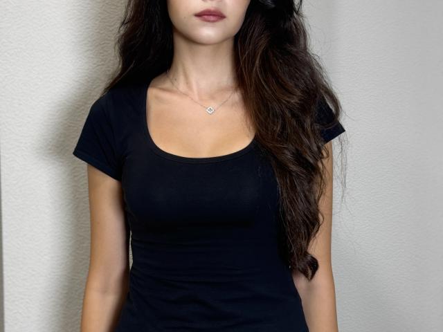 Karoline Valenzano mit langem, dunklem, gewelltem Haar, trägt ein eng anliegendes schwarzes T-Shirt, eine kleine Herz-Kette und roten Lippenstift. Sie zeigt ihre Brüste und ihren Hintern und steht vor einer weißen Wand..