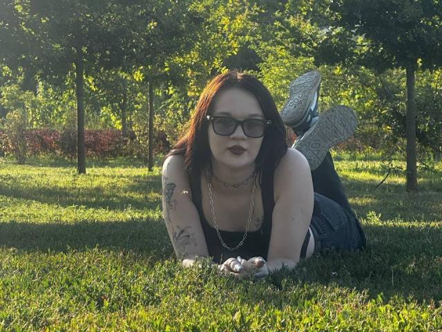 Eliana Laneve deitada na grama, camiseta preta, batom escuro, tatuagens nos braços, óculos, colar de corrente, jeans azuis, tênis, pernas levantadas, iluminada pelo sol, parque verde, relaxada, sexy, fofa..