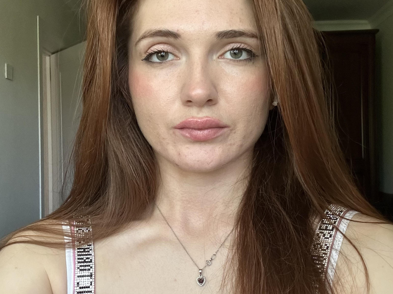 RayeMuck hat langes, braunes Haar, grüne Augen, rosafarbene Lippen, helle Haut und trägt ein weißes Tanktop mit silbernen Trägern. Ihre kleinen Brüste sind leicht sichtbar. Sie schaut in die Kamera..