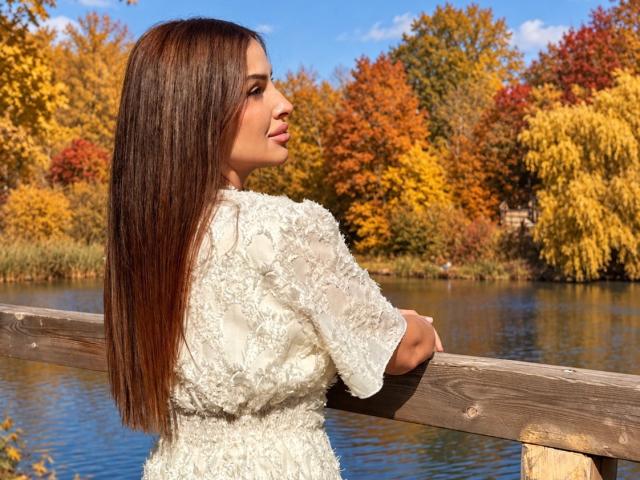 HoneyBlossom with long brown hair, white lace dress, leaning on wooden railing, looking to the side, autumn trees with colorful leaves in the background, bright blue sky.