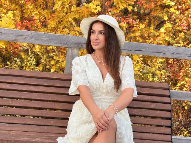 HoneyBlossom sitting on a wooden bench, wearing a white lace dress, hat, and pearl necklace, with hands on her knee, autumn leaves in the background, smiling softly.