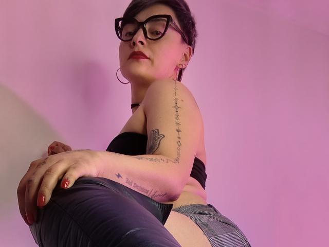 KatParadise with glasses, black strap top, gray plaid pants, tattoos on arm and side, red nail polish, pinkish light, looking down, hand on knee, confident expression.