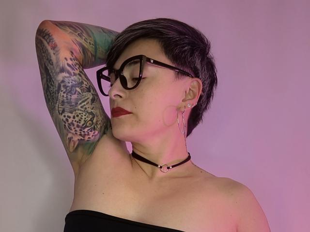 KatParadise with short black hair, glasses, red lipstick, black strapless top, choker, hoop earrings, tattooed arm, posing with arm behind head, pink and purple lighting.