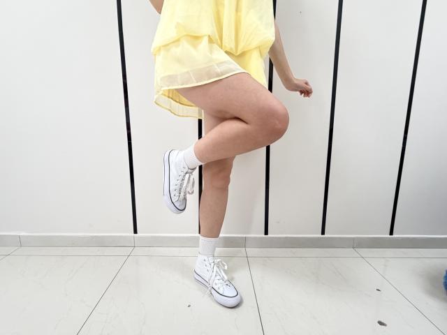 JuliannaMoor's yellow dress, white sneakers, and white socks, standing on one leg with her ass slightly raised, in a minimalist, white-tiled room.