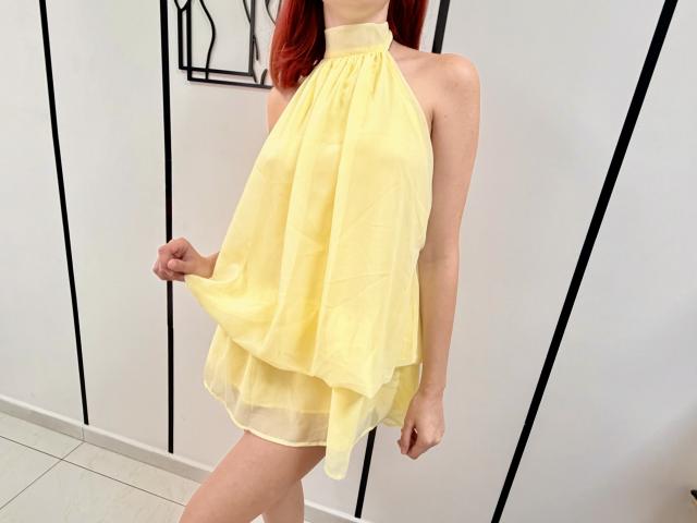JuliannaMoor with red hair, wearing a yellow halter dress, holding it up to show her bare ass, in a bright room.
