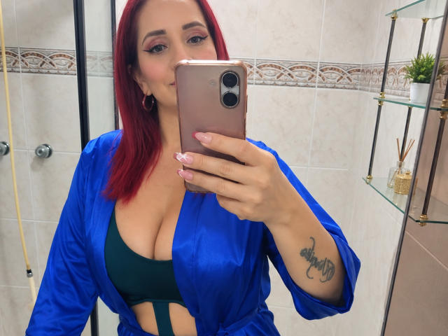 KateMonroe with red hair, wearing a blue satin robe, green bra, showing her tits, holding a pink phone, tattoo on her right forearm, in a bathroom.