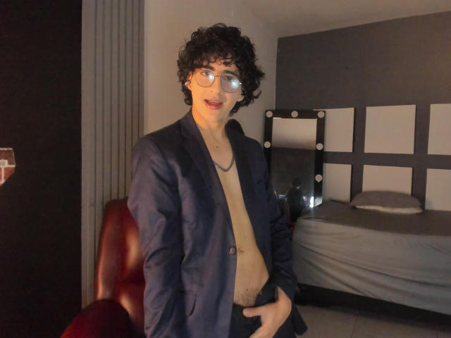 ElliotNoir, curly-haired dude with glasses, open blue jacket, no shirt, hands in black pants, standing in dim room, bed with gray sheets in background, looking at camera.