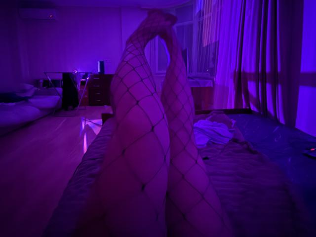 MissKuromi's legs in fishnet stockings, raised and crossed, showing her bare pussy, in a purple-lit bedroom with a bed and dresser in the background.