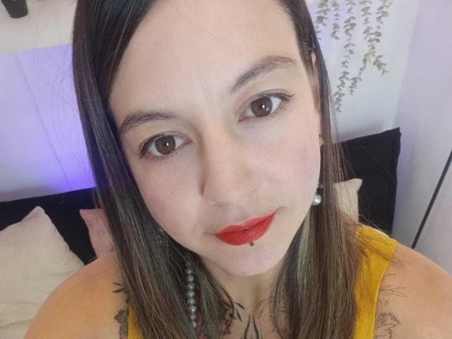 ArianaBlonde has light skin, brown eyes, red lipstick, and a pierced lower lip. She's wearing a yellow top, pearl earrings, and has tattoos on her chest.