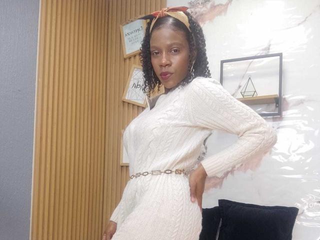 SarahiPusey with curly black hair, wearing a white cable-knit dress, brown belt, and orange headband, standing with hands on hips, black pillow in background.