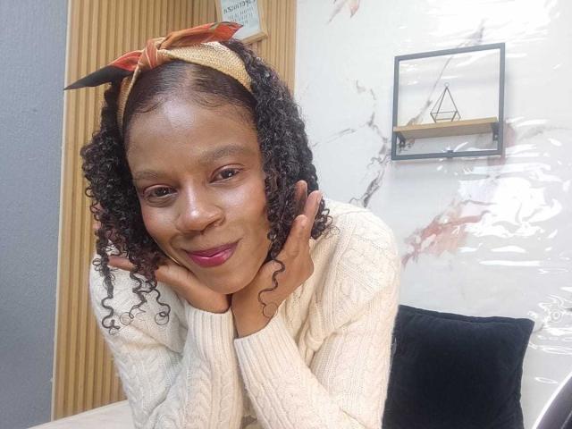 SarahiPusey with curly black hair, brown skin, pink lipstick, wearing a white knit sweater and brown headband, resting her chin on her hands, smiling.