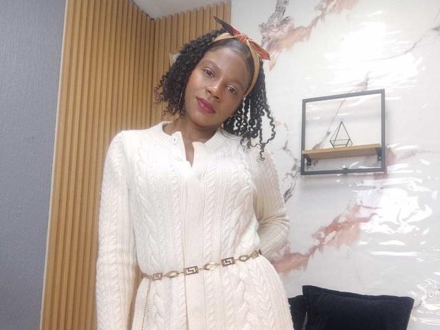 SarahiPusey with curly black hair, wearing a white textured dress with a gold belt, and a pink headband, smiling in a modern room. Her tits are small.