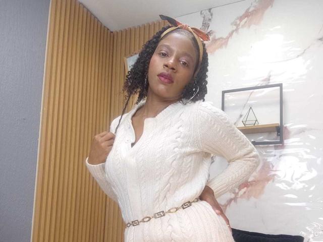 SarahiPusey with curly black hair, wearing a white knitted sweater, orange headband, and belt, standing with hand on hip, showing her ass in a room with wood panels.